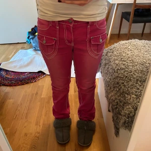 Rosa jeansbyxor med vita sömmar - Säljer ett par riktigt snygga rosa jeansbyxor med kontrasterande vita sömmar och coola fickdetaljer både fram och bak. Byxorna har normal passform och klassisk jeanslook med knapp och dragkedja. Perfekta för dig som vill sticka ut lite extra!
