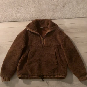 Jacka - Mysig brun teddyjacka från Varley med hög krage och half zip. Jackan har dragsko nertill, långa ärmar och stora fickor framtill. Perfekt för kyliga dagar när du vill vara både varm och snygg.