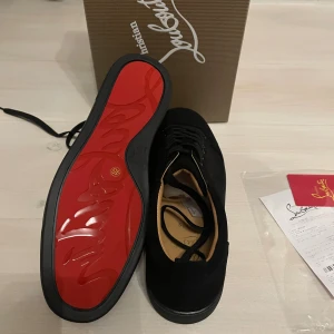 Christian Louboutin svarta sneakers - Säljer ett par svarta sneakers från Christian Louboutin i mocka med klassisk röd sula. Skorna har rund tå, svarta skosnören och diskreta metalldetaljer vid snörningen. Insidan är beige och sulan har Louboutins signaturmönster. Riktigt stilrena och exklusiva sneakers.