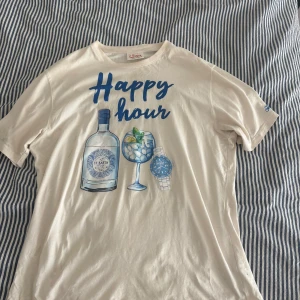 Vit t-shirt MC2 Saint Barth Happy Hour - Snygg vit t-shirt från MC2 Saint Barth i ekologisk bomull. Framsidan har ett stort tryck med texten 'Happy hour', en flaska, ett glas och en blå klocka. T-shirten har rund hals och korta ärmar, perfekt för en chill och avslappnad stil.