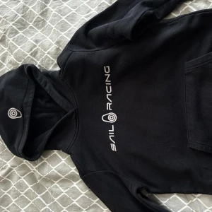 Svart Sail Racing hoodie med tryck - Svart hoodie från Sail Racing med vit logga och tryck på bröstet samt på huvan. Klassisk känguruficka framtill och mjuk insida. Perfekt för dig som gillar sportig och stilren look.