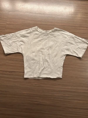 Vit croppad t-shirt från Kappahl - Enkel vit croppad t-shirt från Kappahl med halv långa ärmar och rund hals. Tillverkad i mjuk bomull som känns skön mot huden. Perfekt till jeans eller shorts för en clean och avslappnad look.