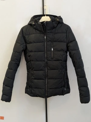 Ridjacka, JH Collection  - Ridjackan har använts en enda gång. Snygg svart ridjacka, vinterjacka, pufferjacka med huva och dragkedja framtill. Jackan har två sidofickor med dragkedja och en bröstficka med dragkedja. Perfekt ridjacka för kyliga dagar och enkel att matcha med olika outfits.