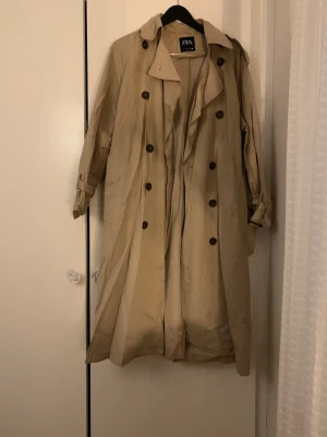 Beige trenchcoat från ZARA - Stilren beige trenchcoat från ZARA med dubbelknäppning och klassisk krage. Jackan har bälte i midjan, knappar vid ärmslut och axelklaffar. Perfekt för lager-på-lager under vår och höst.