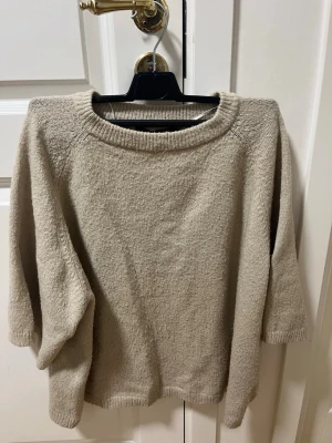 Beige stickad tröja med trekvartsärm - Mysig beige stickad tröja med trekvartsärm och rundad halsringning. Tröjan har en enkel och avslappnad passform med ribbade detaljer vid hals, ärmslut och nederkant. Perfekt för lager på lager under kyliga dagar.