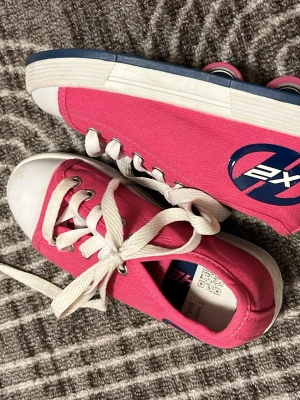Rosa Heelys rollerskor strl 31 - Säljer ett par rosa Heelys rollerskor med vita snören och vit tå. Skorna har blå detaljer och inbyggda hjul i sulan, perfekt för att rulla fram. Ovandelen är i slitstark textil och sulan är blå med mönster för extra grepp. Snygg logga på sidan.