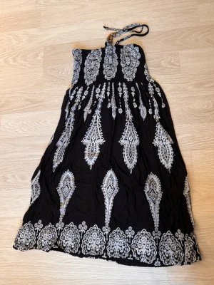 Svart och vit omlottklänning med mönster - Snygg svart omlottklänning med vita detaljerade mönster och knytband i nacken. Klänningen har en bohemisk vibe och är axelbandslös, perfekt för varma dagar. Materialet känns lätt och luftigt, vilket gör den skön att bära.