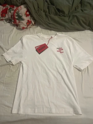 Vit t-shirt från HUGO med tryck - Vit t-shirt från HUGO med rund hals och korta ärmar. På bröstet finns ett tryck i rött och svart med texten 'HUGO YOUR OWN WAY'. T-shirten är i mjuk bomull och har en clean, stilren look som passar perfekt till jeans eller shorts.