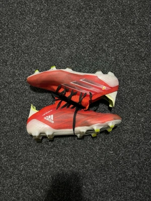 Adidas spedflow .1 - Säljer ett par röda adidas fotbollsskor med svarta snören och vita detaljer. Skorna har en låg profil och platt sula med dobbar i gult och vitt, perfekt för gräsplan. Insidan är ljusgul och ovandelen är tillverkad i syntetmaterial för lätt känsla och bra grepp.