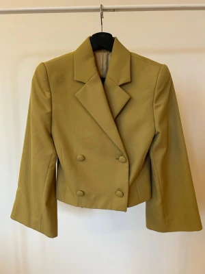 Beige/grön croppad kavaj från TOTEME - Kavaj från TOTEME i beige/grön med dubbelknäppning och breda slag. Jackan har långa ärmar och har satin liknande innan foder. Kavajen är från början en lång kavaj som är uppsydd se sista bilder på hur den är sydd. Inget man se utan är bara på insidan av kavajen.