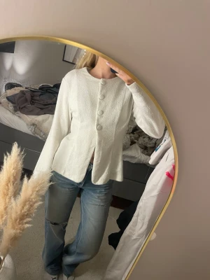 Vit kofta med stora knappar - Säljer en vit kofta med lång ärm och rund hals. Koftan har en lätt struktur i tyget och fyra stora tygklädda knappar framtill. Modellen är figurnära upptill och lite utsvängd nedtill, vilket ger en snygg siluett. Perfekt att styla med jeans eller kjol.