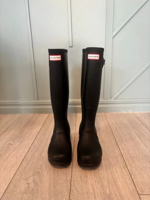 Svarta höga stövlar från Hunter - Säljer ett par klassiska svarta höga Hunter-stövlar i gummi med rund tå och grov sula. De har det ikoniska Hunter-märket framtill och är perfekta för regniga dagar. Insidan har ett ljust mönster och stövlarna är riktigt tåliga och stilrena.