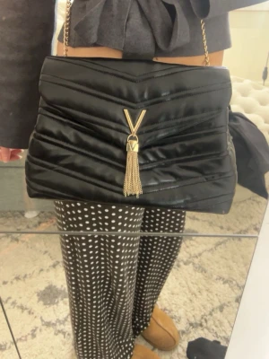 Svart axelväska från Valentino - Säljer en svart axelväska från Valentino med guldfärgad kedja och V-logga framtill. Väskan är helt ny och har aldrig använts, den har till och med plastskyddet kvar kvar på spännet🫶🏻 Nyköpt för 1799kr på Åhléns. Skriv gärna vid frågor