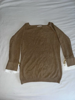 Beige stickad tröja med volangärm - Säljer en beige stickad tröja från NA-KD med långa ärmar och unika detaljer. Ärmsluten har vita plisserade volanger och en liten guldfärgad rosett på varje sida. Perfekt för dig som vill ha något extra till din outfit. STORLEK S/M