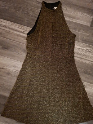 Glittrig svart och guld kortklänning H&M - Supersnygg kort klänning från H&M Divided i svart tyg med glittriga guldtrådar. Ärmlös modell med hög hals och lätt utställd kjol. Perfekt för dig som vill sticka ut med lite extra glitter. Storlek 40.