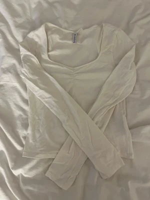 Vit långärmad topp från H&M Divided - Säljer en vit långärmad topp från H&M Divided med snygg v-ringad hals och lätt rynkning framtill. Modellen är figurnära och tillverkad i mjuk bomull, perfekt för att styla med jeans eller kjol. Enkel och clean design som funkar året runt.