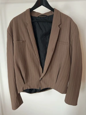  Kort brun blazer i trendig pinstripe – chic & minimalistisk - Brun kavaj med tunna vertikala ränder och enkel knäppning framtill. Jackan har två bröstfickor och är helfodrad med insydd två veck där framme och två där bak. Modellen är lite croppad och har långärmade ärmar. Storlek 52 med axelbredd 50, längd 55. Är en M och haft som lite o-sized kavaj.