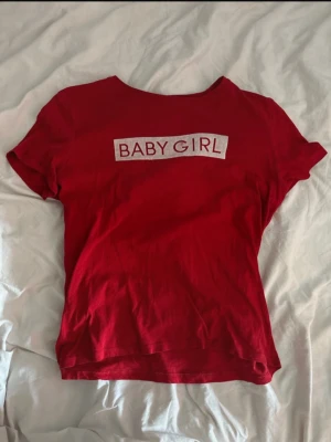 Röd BABY GIRL t-shirt Linn Ahlborg x NA-KD - Säljer en röd t-shirt från Linn Ahlborg x NA-KD i storlek M. T-shirten har korta ärmar och ett vitt tryck med texten 'BABY GIRL' på bröstet. Trycket är lite suddigt