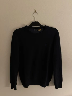 Mörkblå stickad tröja från Polo Ralph Lauren - Snygg mörkblå stickad tröja från Polo Ralph Lauren med diskret logga på bröstet. Rund hals och långa ärmar, perfekt för lager-på-lager. Tröjan har en klassisk passform och ribbade muddar vid ärmslut och nederkant. Passar dig som gillar stilrena och tidlösa plagg.
