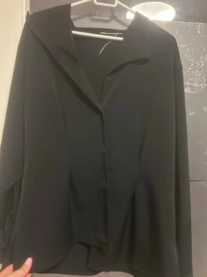 Svart skjorta med klassisk krage - Snygg svart skjorta med klassisk krage och knappar framtill. Skjortan har lång ärm och en stilren, loose passform som ger en chill vibe. Perfekt att matcha med jeans eller kjol för en enkel och cool look.