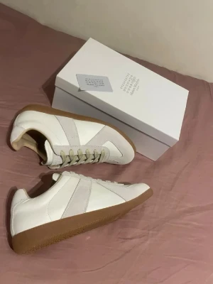 Maison Margiela vita sneakers - Säljer ett par stilrena vita sneakers från Maison Margiela med beige detaljer och gummisula. Skorna har snörning och är tillverkade i skinn och mocka, vilket ger en lyxig känsla. Perfekta för dig som gillar minimalistisk design och neutrala färger.