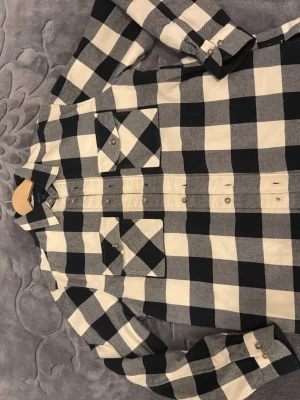 Rutig overshirt jacka från GANT - Säljer en snygg rutig overshirt jacka från GANT i svart och beige. Jackan har quiltat foder, två bröstfickor med knappar och klassisk krage. Materialet är bomull och polyester, perfekt för lager-på-lager under höst och vår. Regular fit och långärmad.