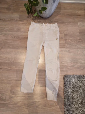 Beige mjukisbyxor från Lyle & Scott - Snygga beige mjukisbyxor från Lyle & Scott med klassisk gul logga på framsidan. Byxorna har elastisk midja med snörning, ribbade muddar vid benslut och en bakficka. Perfekta för att chilla hemma eller träning, tillverkade i mjukt bomullsmaterial. Byxan är 100cm lång, och midjan är 40cm ensidig men går att justeras eftersom de har snören att dra åt och är gjort av bomullsmaterial som gör att den kan stretchas ut medans man har på de några centimeter!✨️