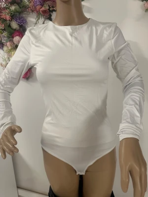 Vit långärmad bodytopp - Säljer en stilren vit bodytopp med lång ärm och rund halsringning. Toppen är figurnära och har en slät, enkel design som passar till många olika looks. Materialet känns mjukt och stretchigt, vilket gör den bekväm att bära hela dagen.