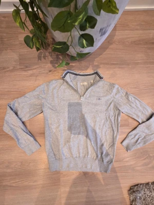 Grå half zip tröja från Gant - Säljer en stilren grå tröja från Gant med half zip och ribbade muddar. Tröjan har en klassisk krage med svarta och vita ränder samt diskret Gant-logo på bröstet. Perfekt till både jeans och chinos för en clean look✨️
