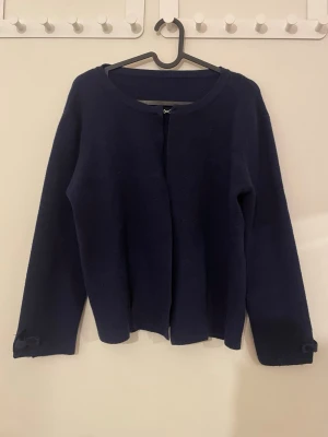 Mörkblå kofta med knappar och hake - Säljer en stilren mörkblå kofta med lång ärm, tillverkad i mjukt stickat material. Koftan har en enkel hake framtill för stängning och dekorativa knappar vid ärmsluten. Perfekt för lager-på-lager under kyliga dagar.