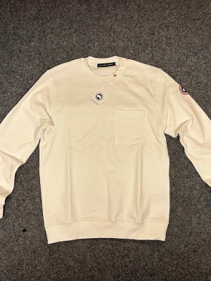 Vit sweatshirt från Canada Goose - Säljer en stilren vit sweatshirt från Canada Goose med rund hals och en bröstficka. Tröjan har lång ärm och klassisk logotyp på vänster ärm. Mjuk bomullskvalitet som känns skön mot huden. Perfekt för dig som gillar enkel och clean design.