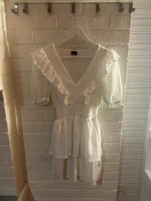 Vit volangklänning från Gina Tricot - Supersöt vit kortklänning från Gina Tricot i storlek XS. Klänningen har puffärmar, v-ringning och volangdetaljer längs axlarna och halsen. Smockad midja och lätt, strukturerat tyg i bomull ger en skön och luftig känsla. Perfekt för sommarens alla tillfällen!