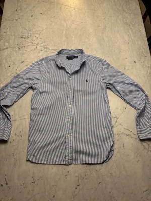 Blårandig skjorta från Ralph Lauren - Snygg blårandig skjorta från Polo Ralph Lauren i slim fit-modell. Skjortan har klassisk krage, långa ärmar och knappar framtill. Tillverkad i mjuk bomull och har loggan broderad på bröstet. Skjortan är 65cm lång och det står att skjortan är i storlek 18 vilket är ca storlek S. Skriv om du har några frågor!
