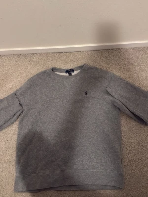 Grå sweatshirt från Polo Ralph Lauren XL - Snygg grå sweatshirt från Polo Ralph Lauren i storlek XL men är inte säker om den riktigt är det. Tröjan har klassisk rund hals, långärmade ärmar och ett litet broderat logomärke på bröstet. Tillverkad i mjuk bomullsmix som är skön mot huden och passar perfekt för chill dagar.