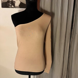 Beige oneshoulder topp från NA-KD - Säljer en beige oneshoulder topp från NA-KD i storlek M. Toppen har en lång ärm på ena sidan och är axelbandslös på andra, vilket ger en cool asymmetrisk look. Materialet är mjukt och stretchigt, perfekt för att styla med jeans eller kjol.
