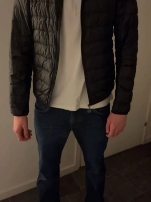 Svart quiltad pufferjacka. EA7 - Svart pufferjacka med quiltad design och dragkedja framtill. Jackan har en stilren look med långärmade ärmar och en normal passform. Perfekt för dig som vill ha en lätt och snygg jacka till kyliga dagar.