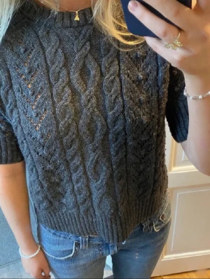 Mörkgrå stickad tröja med kort ärm - Säljer en mörkgrå stickad tröja med korta ärmar och snyggt flätmönster. Tröjan har ribbad kant vid hals, ärmslut och nederkant.
