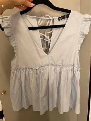 Ljusblå blus med snörning från Zara - Supersöt ljusblå blus från Zara med volangdetaljer på axlarna och v-ringning både fram och bak. Blusen har snörning i ryggen och ett rynkat parti under bysten som ger en flowy känsla. Perfekt för vår och sommar!