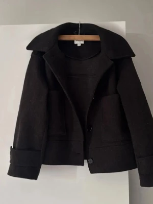 Mörkgrå kort kappa - Säljer en mörkgrå kort kappa med bred krage, stora fickor framtill och knäppning med två knappar. Jackan har en rak, boxig passform med långa ärmar och stilrena detaljer. Perfekt för dig som gillar minimalistisk och clean stil.