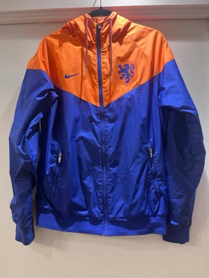 Nike vindjacka KNVB  - Nike windbreaker för Nederländernas fotbollslandslag KNVB. Vindjackan är i orange och blå med KNVB-lejonet på bröstet. Jackan har huva med snörning, dragkedja framtill och två fickor med dragkedja. Storlek S. Mycket bra skick! Originalpris: 1500kr. Säljs på flera sidor, först till kvarn. 