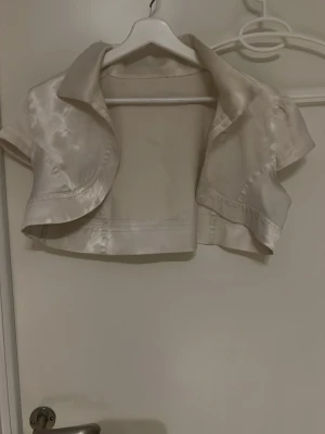 Champagnefärgad kort bolero i satin - Snygg kort bolero i champagnefärgad satin med glansig finish. Modellen har korta ärmar och öppen front, perfekt att slänga över en klänning för en extra touch. Klassisk krage och elegant snitt som ger en chic look.