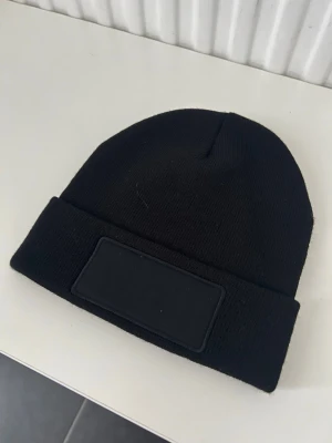 Svart stickad mössa med patch - En stilren svart stickad mössa med uppvikt kant och en svart patch framtill. Mössan har en enkel och clean look som passar till de flesta outfits. Materialet är mjukt och stretchigt för skön passform.