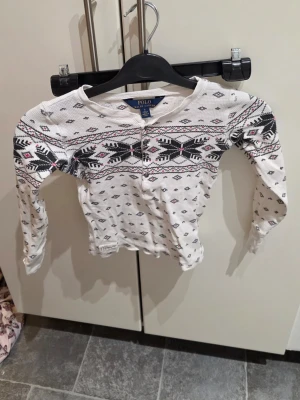 Vit långärmad tröja Polo Ralph Lauren - Vit långärmad tröja från Polo Ralph Lauren med svart och rosa mönster över bröstet och axlarna. Tröjan har rund hals och knappar framtill. Materialet är bomull med en strukturerad yta som ger en mysig känsla. Perfekt för kyliga dagar.