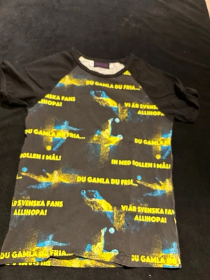 Svart t-shirt Design Roshill, fotbollstryck - Cool svart t-shirt från Design Roshill i storlek 146. Tryck med blå och gula fotbollsspelare och texter som 'Du gamla du fria' och 'Vi är svenska fans allihopa'. Perfekt för dig som älskar fotboll och vill sticka ut med färgstarkt mönster.