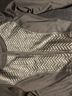 Svart croppad quiltad jacka Made in Italy - Snygg svart croppad jacka med quiltade paneler i ett zigzag-mönster på framsidan. Jackan har långärmade och en cool, modern look som passar perfekt till streetwear. Materialet är polyester och jackan är tillverkad i Italien.