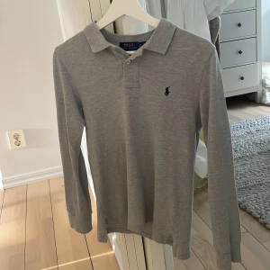 Grå långärmad pikétröja Polo Ralph Lauren - Klassisk grå långärmad pikétröja från Polo Ralph Lauren med broderad marinblå logga på bröstet. Tröjan är i 10-12 år men är stor i storleken, skulle rekommendera tröjan till dig som är 160-170cm! Tröjan är i grymt skick med inga defekter