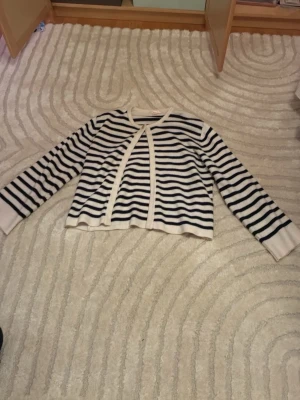 Randig kofta från Gina Tricot - Säljer en snygg vit och marinblå randig kofta från Gina Tricot i storlek 158/164. Koftan har lång ärm, öppen framtill och är stickad i mjukt material. Perfekt för lager på lager och ger en fräsch look till din outfit.