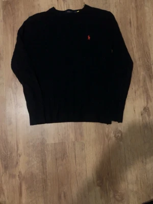 Svart kabelstickad tröja Polo Ralph Lauren - Svart kabelstickad tröja från Polo Ralph Lauren i storlek S. Klassisk design med rund hals och rött broderat logomärke på bröstet. Tröjan har lång ärm och är gjord i mjukt stickat material som känns skönt mot huden. Priset kan diskuteras om inte för lågt.