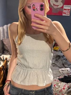 Vit broderad linnetopp från Kappahl - Superfin vit linnetopp från Kappahl med broderade hålmönster och smala axelband. Toppen har resår i midjan som ger en söt volangkant nertill. Perfekt för varma dagar och enkel att matcha med jeans eller kjol.