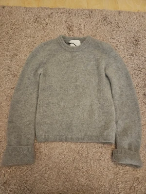 Grå stickad tröja H&M Edition XS - Jag säljer en supermjuk, superfin och fluffig ljusgrå stickad tröja från New Edition. Rund halsringning och långa ärmar med ribbade muddar. Perfekt för att hålla dig varm och snygg under kyliga dagar. Använt endast en gång och i mycket bra skick. Nypris runt 600-700 kr.